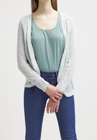 Licht mintgroene gebreide cardigan met een open voorkant, textuurstof en lange mouwen, gelaagd over een teal top en donkerblauwe jeans.