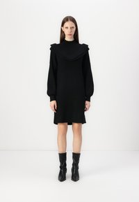 Juoda megzta suknelė su plačiu, gofruotu apykaklė ir ilgomis rankovėmis. Suknelė pasižymi tiesia siluetu ir baigiasi šiek tiek virš kelių.