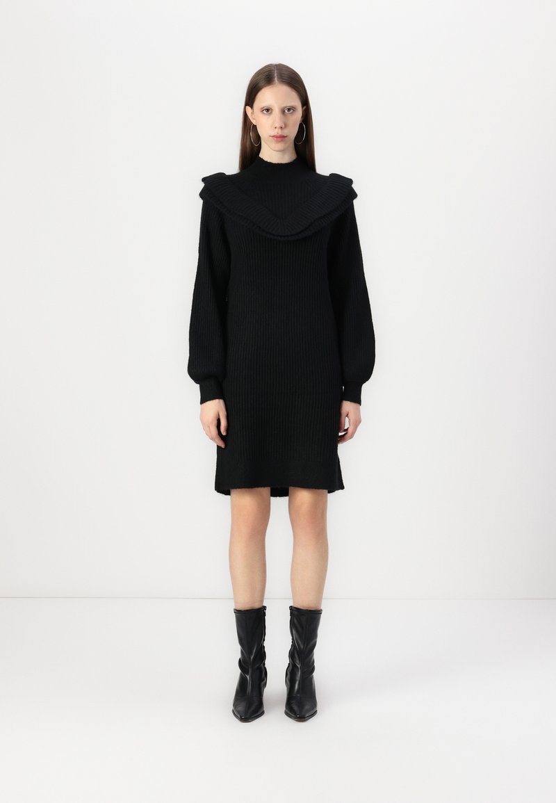 Juoda megzta suknelė su plačiu, gofruotu apykaklė ir ilgomis rankovėmis. Suknelė pasižymi tiesia siluetu ir baigiasi šiek tiek virš kelių.