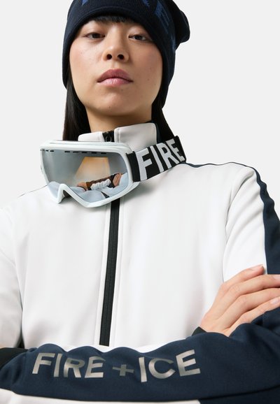 Skijacke in Weiß mit marineblauen Akzenten, mit Reißverschluss und Logo auf dem Ärmel. Skibrille mit weißem Rahmen ruht auf dem Kragen.