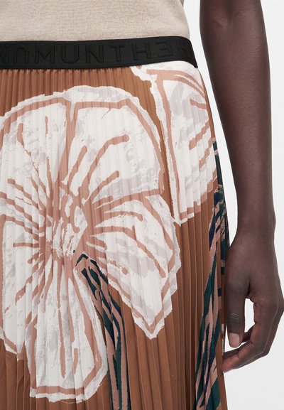 Jupe plissée avec grand imprimé floral blanc et marron, taille noire avec du texte, portée avec un haut beige et un bras à la peau foncée visible sur le côté droit.