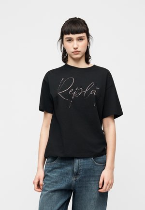 Junge Frau mit kurzen dunklen Haaren, die ein schwarzes, übergroßes T-Shirt mit dem Schriftzug "Replay" und lockere blaue Jeans vor einem schlichten weißen Hintergrund trägt.