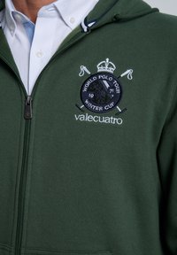 Sudadera verde con cremallera hecha de material suave. Presenta un logo bordado con una corona, palos de polo cruzados y texto que detalla "World Polo Tour".