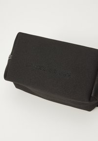 Borsa rettangolare nera realizzata in tessuto texturizzato con logo in rilievo. Presenta una chiusura con zip e un sottile accento di cucitura bianca.