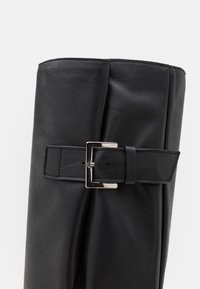 Bota de cuero negro con caña y una hebilla rectangular de plata en una correa ajustable, sobre un fondo blanco liso.