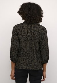 Blouse à fleurs foncées avec des manches trois quarts, en tissu texturé, col à volants et une coupe légèrement ample. Motif noir sur fond vert olive.
