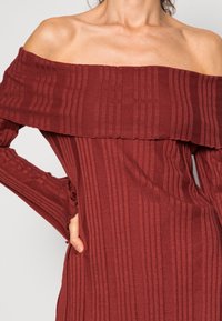 Off-the-shoulder burgundy dress with ribbed texture, long sleeves, and ruffled hem detailing. Form-fitting silhouette enhances the design.