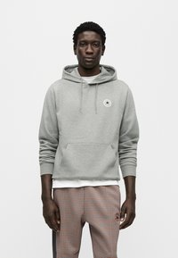 Γκρι φούτερ με κουκούλα, μπροστινή τσέπη και ρυθμιζόμενο κορδόνι. Διαθέτει κυκλικό λογότυπο στο στήθος. Φοριέται πάνω από λευκό πουκάμισο και καρό παντελόνι.