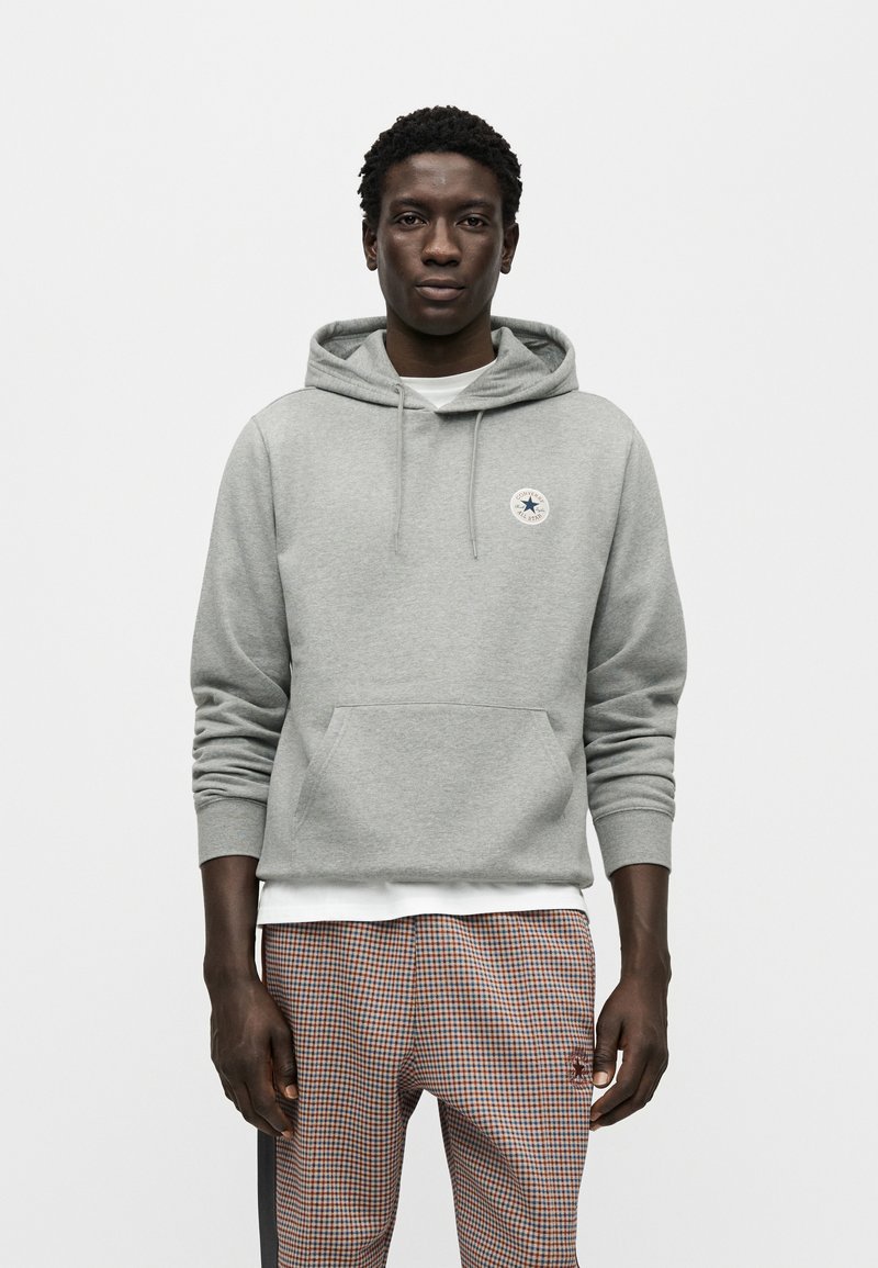 Grå hættetrøje med en forlommelomme og justerbar snøre. Har et rundt logo på brystet. Båret over en hvid skjorte og ternede bukser.