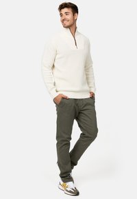 Suéter de punto crema con cremallera de un cuarto y acento marrón, combinado con pantalones verde oliva y zapatillas deportivas grises con detalles amarillos.