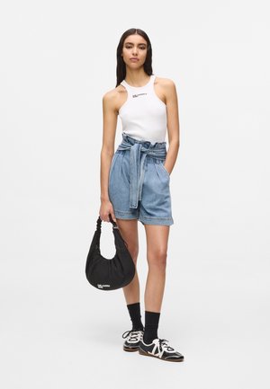 Nő fehér ujjatlan felsőben, magas derekú papírzacskó fazonú farmerrövidnadrágban övvel, fekete zokniban, fekete-fehér tornacipőben, fekete kézitáskát tartva.