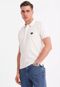 Hvid polo skjorte med et sort emblem, korte ærmer og en krave med tre knapper. Sammen med blå jeans og et sølvur.