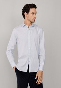 Hombre vistiendo una camisa de cuadros en tonos azul claro con mangas largas, de pie con una mano en el bolsillo, mirando hacia un lado.