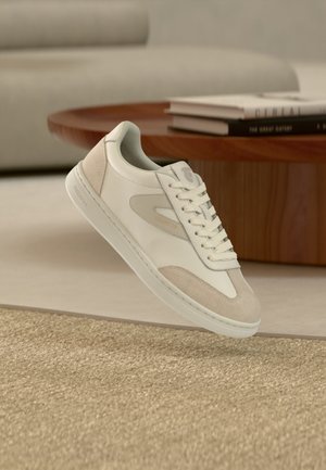 Witte en beige lage sneaker met veters zwevend boven een geweven vloerkleed in een woonkamer met een houten ronde tafel en gestapelde boeken.