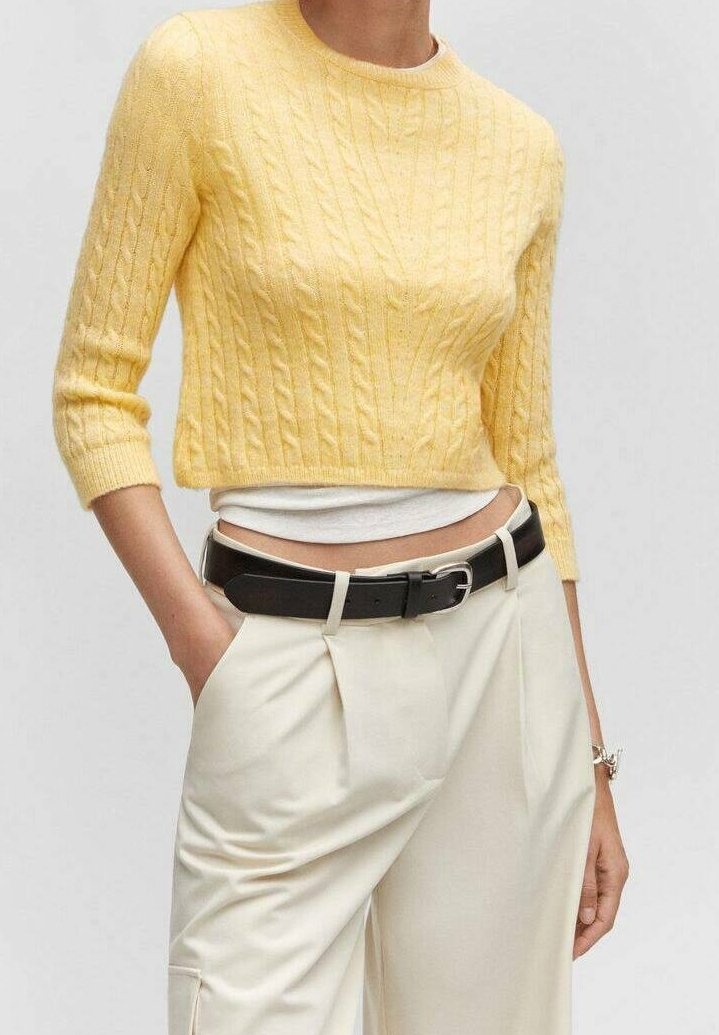 Femme portant un pull court jaune en tricot torsadé par-dessus un haut blanc, un pantalon crème taille haute et une ceinture noire, les mains dans les poches.