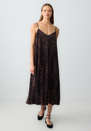 Donna che indossa un vestito lungo fino alle caviglie, senza maniche, ampio, con spalline sottili e scarpe nere con cinturini alla caviglia su uno sfondo chiaro e uniforme.