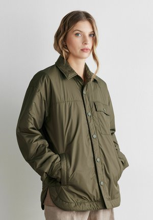 Kvinde med bølget hår iført en oversized olivengrøn skjortejakke med knapper og lommer, stående mod en ensfarvet hvid baggrund.