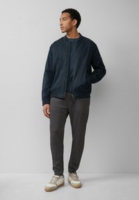 Marine bomberjack met rits, gecombineerd met grijze tappered broeken en crèmekleurige sneakers met beige accenten en textuurdetails.