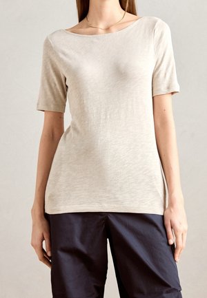 Frau trägt ein beiges Kurzarmoberteil und marineblaue Hose, steht vor einem schlichten hellen Hintergrund und trägt eine dünne goldene Halskette.