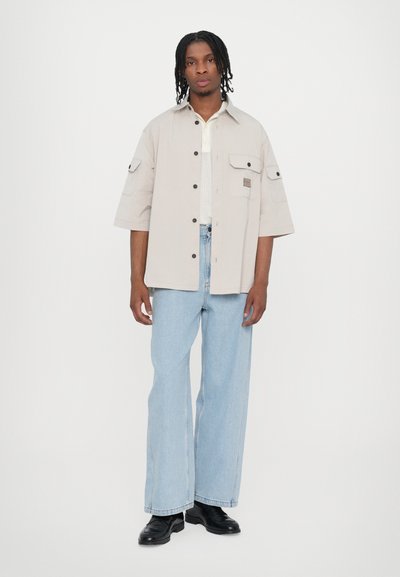 Chemise oversize beige clair à manches courtes, fermeture bouton, et deux poches avant, associée à un jean large bleu clair et des bottes noires.