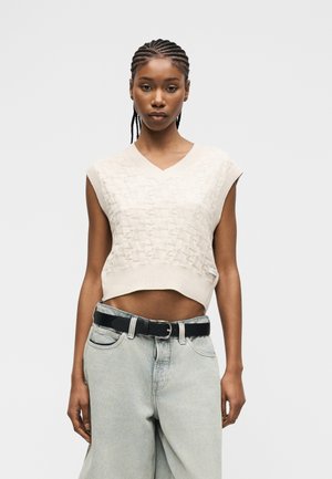 Nő fonott hajjal, bézs színű, ujjatlan kötött crop topot és világoskék, bőszárú farmert fekete övvel viselve, egyszerű háttér előtt áll.