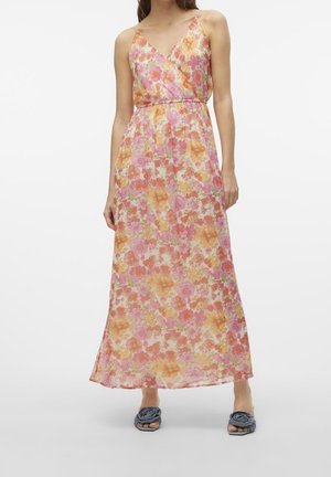 Vrouw die een mouwloze, V-hals, enkellange jurk draagt met een roze, oranje en geel bloemenpatroon, gecombineerd met blauwe sandalen met knoop.