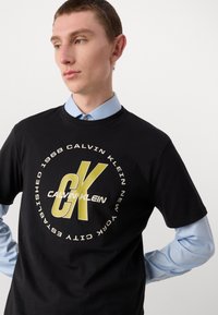Melna kokvilnas T-krekls ar izteiksmīgu dzeltenu un baltu apļveida logo, kurā ir teksts "Calvin Klein". Īsas piedurknes, apaļa kakla izgriezums, gluda tekstūra.