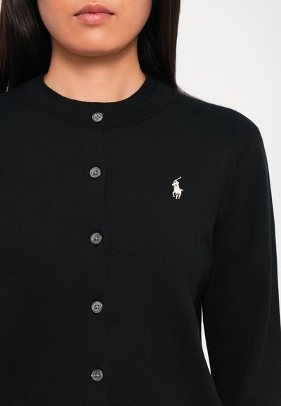 Donna che indossa un cardigan nero con bottoni, con un piccolo logo di un giocatore di polo bianco sul petto sinistro, mostrata dalla zona del collo fino alla parte centrale del torace.