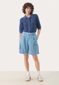 Sininen neulecardigan, jossa on teksturoitu zikzakkikuvio, yhdistettynä vaaleansinisiin raidoitettuihin shortseihin. Mallilla on valkoiset sukat ja lenkkarit.