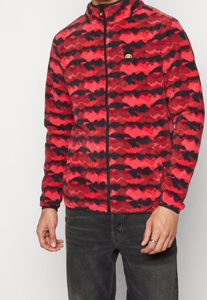 Mann trägt eine rot-schwarze Fleecejacke mit Camouflage-Muster, einem Frontreißverschluss und einem Logo-Patch auf der Brust, kombiniert mit schwarzer Jeans.