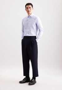 Uomo in piedi con le mani in tasca, che indossa una camicia azzurro chiaro, pantaloni navy tagliati, calzini neri e scarpe eleganti nere.