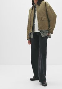 Veste bomber kaki avec col marron, superposée à une chemise à carreaux. Jeans noirs à jambes larges et ballerines noires complètent le look. Design simple.