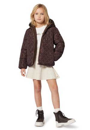 Veste en doudoune matelassée marron avec capuche, présentant un motif et une fermeture éclair. Portée par-dessus une chemise blanche et une jupe plissée claire avec des bottes.