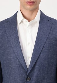 Tamsiai mėlynas tekstūruotas švarkas su suapvalintais atlapais, su vienu juodu mygtuku, vilkimas ant balto marškinėlių su apykakle, kuriame matyti mygtukai.