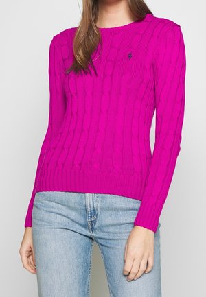 Vrouw draagt een felroze kabelgebreide trui met lange mouwen en lichtblauwe jeans, staand tegen een effen achtergrond.