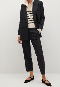 Femme portant un blazer noir à double boutonnage, un pantalon noir raccourci, un pull en maille rayé crème et noir, et des ballerines pointues noires.