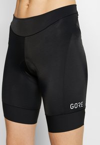 Czarne spodenki rowerowe z rozciągliwego materiału, z szerokim paskiem w talii, wyściełanym obszarem siedzącym oraz małym białym logo po stronie dolnej.