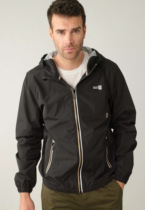 Veste à capuche noire en tissu léger, avec fermeture éclair à l'avant, poignets élastiques et deux poches latérales zippées. Équipée de finitions en beige.