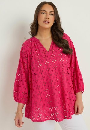 Kobieta w jasnoróżowej bluzce z ażurowym wzorem kwiatowym i białych spodniach, pozująca na tle jednolitego jasnego tła.