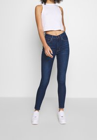 Höga mörkblå slim fit-jeans med en slät textur och diskreta sömdetaljer, i kombination med en kort vit tanktop och vita sneakers.
