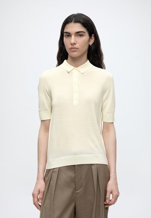 Kvinde med langt mørkt hår iført en cremefarvet kortærmet knappet poloshirt og løse brune bukser, stående mod en ensfarvet hvid baggrund.