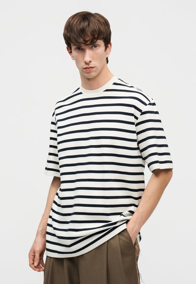 Νέος άνδρας που φοράει κοντομάνικο λευκό και μαύρο πουκάμισο με οριζόντιες ρίγες και καφέ παντελόνι, στέκεται με το ένα χέρι στην τσέπη.