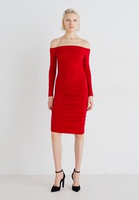 Rochie roșie, cu umerii expuși, cu mâneci lungi, textură plisată, până la jumătatea gambei. Purtați cu tocuri stiletto negre, fundalul este alb simplu.