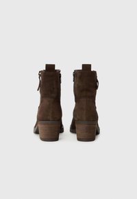Botas de tobillo de ante marrón con tacón apilado, cremallera lateral y tirador. Presenta una textura suave y un diseño de punta redondeada.