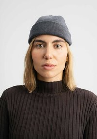 Grijze gebreide muts met een omgeslagen rand, gedragen over een bruin, hoog gesloten ribtrui. De muts heeft een gestructureerd oppervlak en een subtiel patroon.