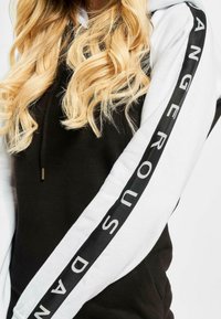 Crna i bijela hoodie sa dugim rukavima od kontrastnog materijala. Jedan rukav ima vertikalnu crnu traku s natpisom "DANGEROUS" u bijelim slovima.