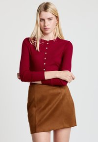 Langärmliges, geripptes Oberteil in Burgunderrot mit rundem Ausschnitt und Knopfdetails, kombiniert mit einem braunen Minirock aus Wildleder.
