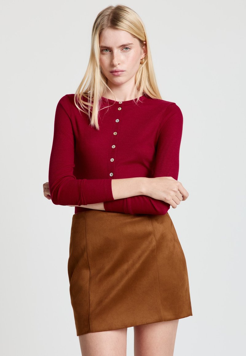 Langärmliges, geripptes Oberteil in Burgunderrot mit rundem Ausschnitt und Knopfdetails, kombiniert mit einem braunen Minirock aus Wildleder.