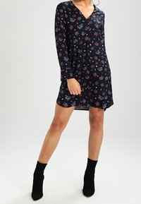 Námořnické modré květinové šaty s dlouhými rukávy a výstřihem do V. Mají malé červené a bílé vzory. V kombinaci s černými kotníkovými botami.
