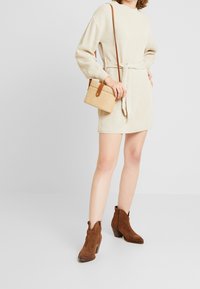 Beige texturerad klänning med bälte i midjan, tillsammans med en liten vävd handväska och bruna mocka ankelstövlar.